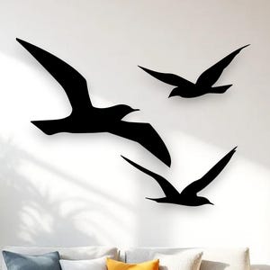 Puede incluir: Tres siluetas de aves negras montadas en una pared. El ave m&aacute;s grande est&aacute; situada a la izquierda con las alas extendidas, mientras que dos aves m&aacute;s peque&ntilde;as est&aacute;n dispuestas a la derecha, una ligeramente m&aacute;s alta, todas en pleno vuelo.