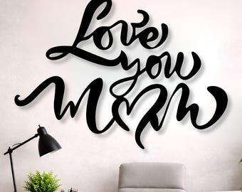 Puede incluir: Arte de pared de metal negro con las palabras "Love you Mom" en una fuente cursiva. La obra de arte está montada en una pared blanca sobre un escritorio con una computadora portátil, una lámpara y una planta en maceta.