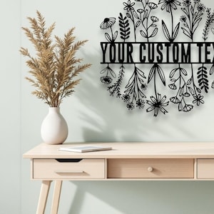 Puede incluir: Arte mural floral de metal negro con el texto "YOUR CUSTOM TEXT" en un dise&ntilde;o circular. La obra de arte presenta varias siluetas de flores y hojas. La pieza se exhibe en una pared verde claro sobre un escritorio de madera.