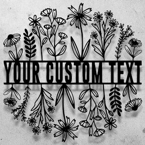 May include: Black metal floral wreath with the text "YOUR CUSTOM TEXT" across the center. The wreath features various stylized flowers and foliage, creating a decorative design. The background is a textured, light gray surface.