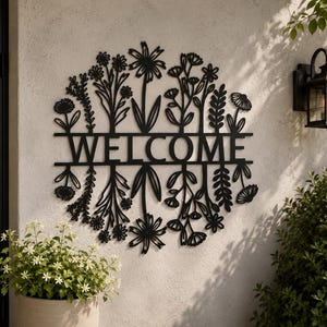 May include: Black metal WELCOME sign featuring a floral design. The sign is mounted on a textured wall, with a black lantern and greenery in the background. The word WELCOME is cut out in large, bold letters.
