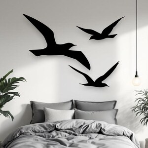 Puede incluir: Tres siluetas de aves negras en vuelo est&aacute;n montadas en una pared lisa sobre una cama. Las aves se representan en diferentes posiciones, con un ave grande a la izquierda y dos aves m&aacute;s peque&ntilde;as a la derecha, creando una escena din&aacute;mica.