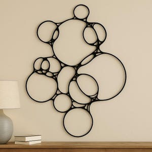 May include: Black, abstract wall art featuring interconnected circular shapes of varying sizes. The design is mounted on a neutral-colored wall, creating a modern and minimalist aesthetic. The artwork is a decorative piece for home decor.