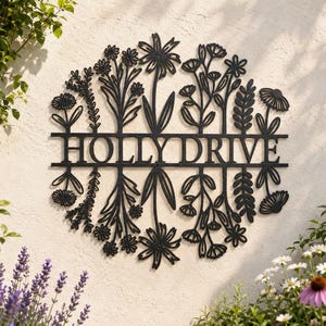 May include: Black metal sign reading "HOLLY DRIVE" encircled by a floral arrangement. The sign is fixed to a light-coloured textured wall, with plants and flowers in the foreground.