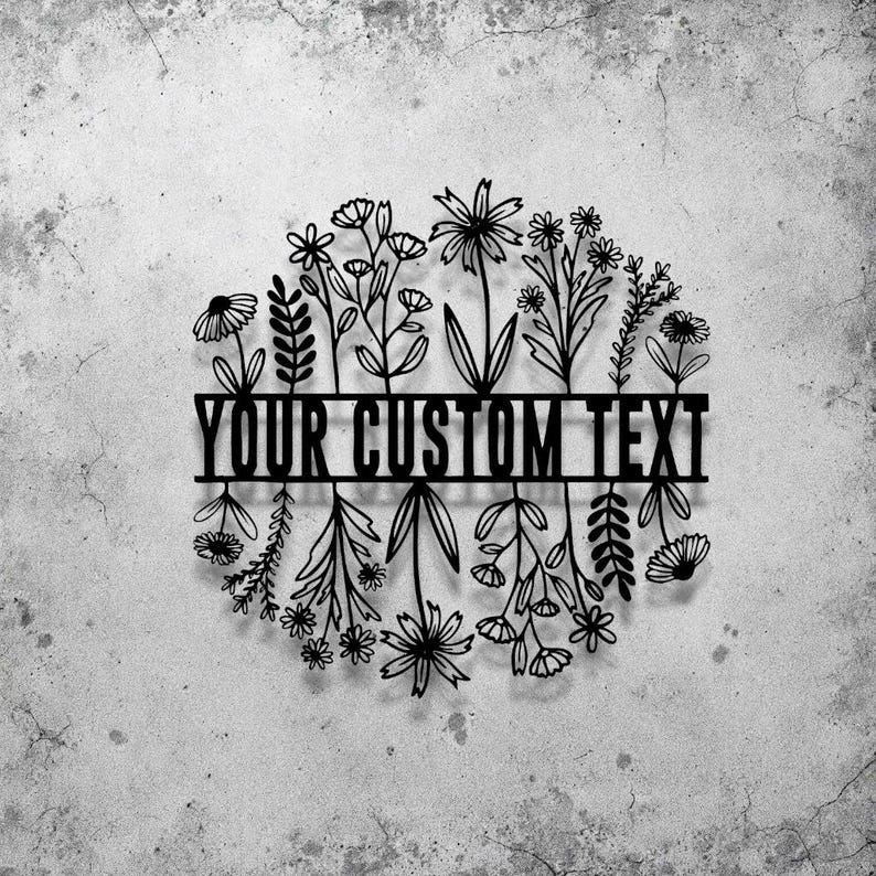 May include: Black metal floral wreath with the text "YOUR CUSTOM TEXT" across the center. The wreath features various stylized flowers and foliage, creating a decorative design. The background is a textured, light gray surface.