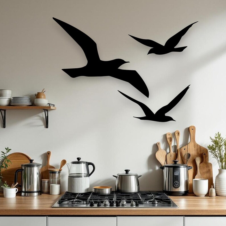Puede incluir: Tres siluetas de aves negras est&aacute;n montadas en una pared sobre una encimera de cocina. El arte de pared presenta un ave grande con las alas extendidas y dos aves m&aacute;s peque&ntilde;as en vuelo. La cocina incluye una estufa, ollas y tablas de madera.