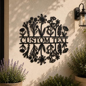 May include: Black metal floral wall art with the words "CUSTOM TEXT" in the centre. The art features various flower and leaf designs. The art is mounted on a beige wall, with a black light fixture and plants in the background.