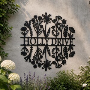 May include: Black metal sign featuring the text "HOLLYDRIVE" within a floral wreath design. The sign is mounted on a light grey wall, with plants and flowers in the foreground.
