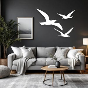 Puede incluir: Tres siluetas de aves blancas montadas en la pared, dispuestas en formaci&oacute;n de vuelo sobre una pared gris oscuro. Las aves est&aacute;n representadas en un estilo minimalista con las alas extendidas, creando un efecto visual din&aacute;mico sobre un sof&aacute; gris.