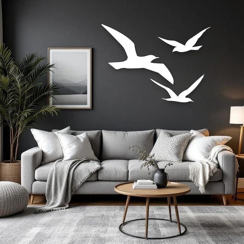 Puede incluir: Tres siluetas de aves blancas montadas en la pared, dispuestas en formaci&oacute;n de vuelo sobre una pared gris oscuro. Las aves est&aacute;n representadas en un estilo minimalista con las alas extendidas, creando un efecto visual din&aacute;mico sobre un sof&aacute; gris.
