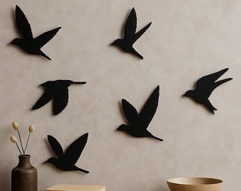 May include: Five black bird silhouettes are mounted on a wall in a flying formation. Below them, a dark brown vase holds dried stems, a stack of two light-colored books sits on a wooden surface, and a beige bowl rests to the right side.