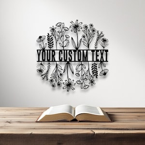 Puede incluir: Arte mural floral de metal negro con el texto "YOUR CUSTOM TEXT" en una barra horizontal. El arte presenta una disposici&oacute;n circular de flores y follaje estilizados. Un libro abierto descansa sobre una mesa de madera.