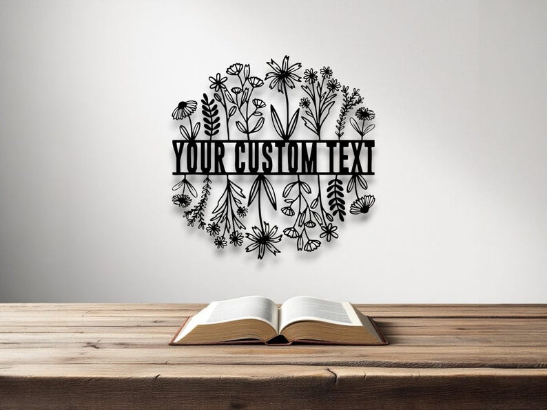 Puede incluir: Arte mural floral de metal negro con el texto "YOUR CUSTOM TEXT" en una barra horizontal. El arte presenta una disposici&oacute;n circular de flores y follaje estilizados. Un libro abierto descansa sobre una mesa de madera.