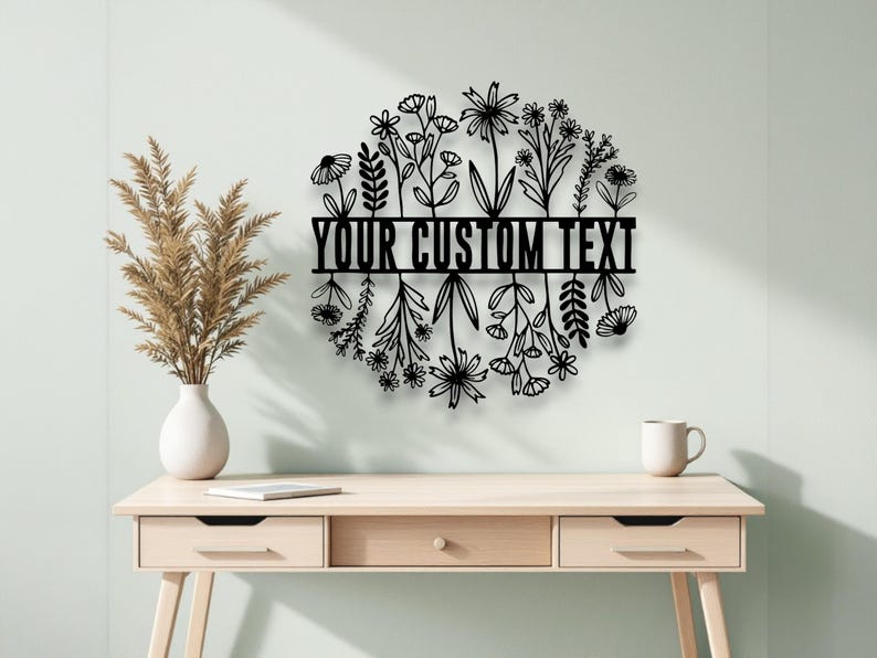 Puede incluir: Arte mural floral de metal negro con el texto "YOUR CUSTOM TEXT" en un dise&ntilde;o circular. La obra de arte presenta varias siluetas de flores y hojas. La pieza se exhibe en una pared verde claro sobre un escritorio de madera.