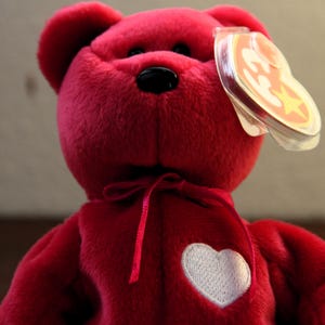 Può includere: Un orsacchiotto di peluche rosso con un cuore bianco sul petto. L'orso indossa un nastro rosso intorno al collo e un berretto rosso con una stella bianca.