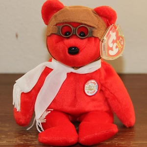 Puede incluir: Oso de peluche rojo de la marca Beanie Baby con un gorro de aviador marrón con gafas y una bufanda blanca. El oso tiene una nariz negra y ojos negros. El oso tiene una etiqueta circular que dice "Ty" y un parche circular que dice "100 Years of Flight".