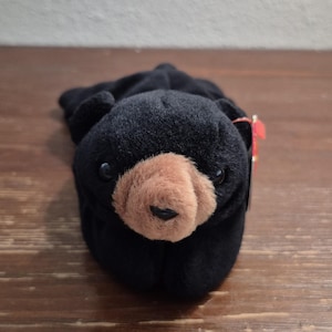 May include: A black plush bear toy with a brown snout and small black eyes. The bear is lying down on a wooden surface. A red tag is attached to the bear's ear.