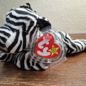 Peut inclure: Un jouet en peluche zèbre rayé noir et blanc avec une étiquette Ty Beanie Baby rouge et jaune. L'étiquette indique "Beanie Original Baby".