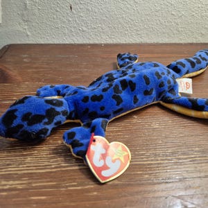 Puede incluir: Un juguete de peluche de lagarto azul con manchas negras y una lengua roja. El juguete tiene una barriga y pies amarillos. Una etiqueta en forma de corazón está unida al juguete.