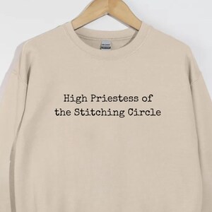 May include: A beige sweatshirt with the text "High Priestess of the Stitching Circle" in black. The sweatshirt is hanging on a wooden hanger against a white background. The long-sleeved top has a crew neck.