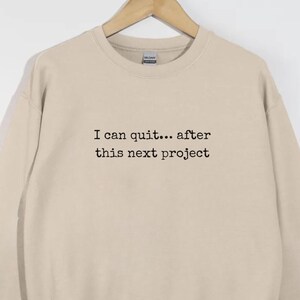 May include: A beige sweatshirt with the text "I can quit... after this next project" in black. The sweatshirt is hanging on a wooden hanger. The garment has a crew neck and long sleeves.