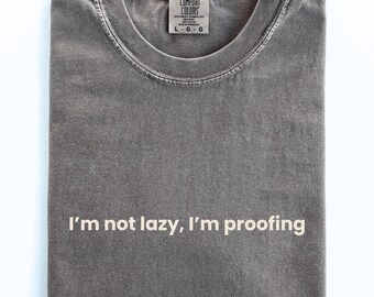 May include: A gray Comfort Colors t-shirt, folded neatly, with the text "I'm not lazy, I'm proofing" in white. The shirt has a crew neck and a tag at the neckline.