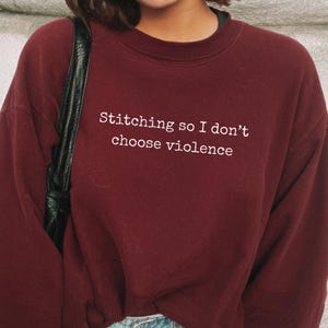 May include: A maroon sweatshirt with the text "Stitching so I don't choose violence" in white. The sweatshirt is a crew neck style and appears to be made of a soft material. The text is centered on the front of the shirt.