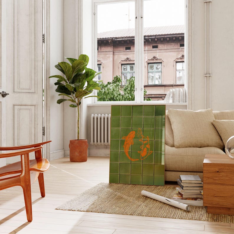 May include: A green tile art piece featuring an orange koi fish design. The artwork is leaning against a beige sofa in a well-lit room with a large window. A potted plant and wooden chair are also visible.