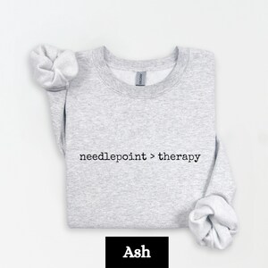 May include: Light gray sweatshirt with the text "needlepoint > therapy" in black. The sweatshirt is folded, with the sleeves rolled up. The word "Ash" is in a black rectangle at the bottom.