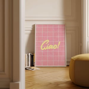 May include: A framed pink grid art print with the word "Ciao!" in yellow script. The artwork is leaning against a wall, next to a stack of books and a yellow ottoman. The floor is made of wood.