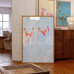 May include: A framed print featuring a watercolor illustration of an orange bikini hanging on a clothesline against a blue sky background. Additional framed prints depict a pink scooter and palm trees with "Palm Springs" text.