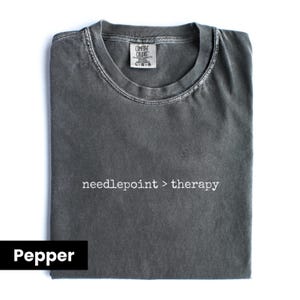 May include: A pepper-colored, short-sleeved t-shirt with the text "needlepoint > therapy" printed in white. The shirt has a crew neck and a tag at the back of the neck. The shirt is folded neatly on a white background.