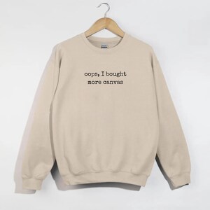 May include: A beige sweatshirt with the text "oops, I bought more canvas" in black. The sweatshirt is hanging on a wooden hanger. The garment has long sleeves and a crew neck.