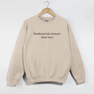 May include: A beige sweatshirt with the text "Needlepoint: cheaper than bail" in black. The sweatshirt is hanging on a wooden hanger against a white background. The garment has a crew neck and long sleeves.