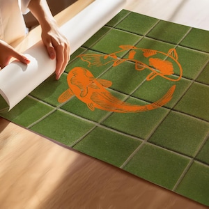 May include: Green tile-patterned wallpaper with two orange koi fish illustrations. The wallpaper is being unrolled on a wooden surface. The koi fish are in a circular design.