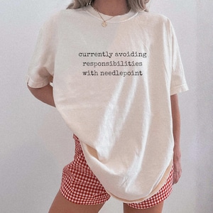 May include: Cream-colored t-shirt with the text "currently avoiding responsibilities with needlepoint" in black. The person is wearing red and white checkered shorts. The shirt has short sleeves and a crew neck.