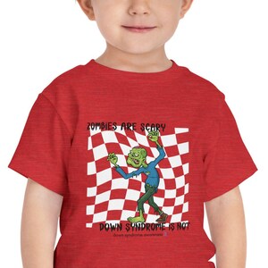 Puede incluir: Una camiseta roja para niños pequeños con un diseño de zombi de dibujos animados sobre un fondo a cuadros. El zombi es verde con pantalones azules y levanta los brazos. El texto "Zombies are scary, Down Syndrome is not" está impreso en la camiseta.