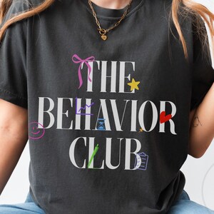 Puede incluir: Camiseta gris oscuro con el texto blanco "THE BEHAVIOR CLUB". El diseño incluye un lazo rosa, una estrella amarilla, un corazón rojo y otros gráficos coloridos. La camiseta es de un material suave.