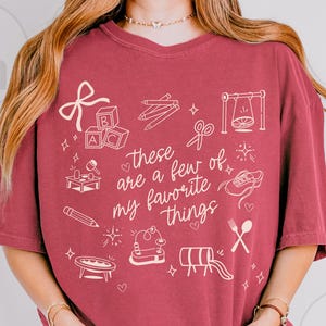 May include: Burgundy-colored t-shirt with the phrase "these are a few of my favorite things" and various hand-drawn illustrations. The illustrations include a bow, alphabet blocks, pencils, scissors, a swing, and other playful objects.