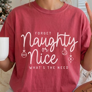 Op de afbeelding: Een rood t-shirt met korte mouwen en witte tekst die luidt Forget Naughty or Nice WHAT'S THE NEED. Het ontwerp bevat witte lijntekeningen van drie hangende kerstballen die tussen de woorden Naughty en Nice zijn geplaatst.