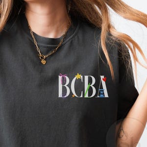 以下が含まれることがあります： 白い「BCBA」の文字がカラフルなグラフィックで飾られた、濃い灰色のTシャツ。 Tシャツはクルーネックと半袖です。 ゴールドのネックレスが見えます。