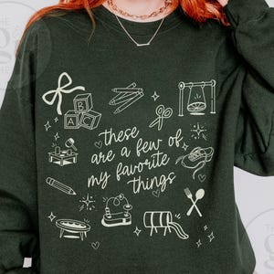 May include: Dark green sweatshirt with white line art illustrations of toys and the phrase "these are a few of my favorite things." The design includes blocks, pencils, scissors, a swing, and a shoe.