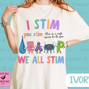 Op de afbeelding: Een ivoorkleurig t-shirt met de kleurrijke tekst I STIM you stim this is a safe space to be you WE ALL STIM. De afbeelding bevat cartoonillustraties van een blauwe sensorische schommel, een roze pop-it, een paarse fidget spinner, een trampoline en een groene letter P.