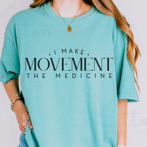 May include: A turquoise short-sleeved t-shirt with the text "I MAKE MOVEMENT THE MEDICINE" in black. The shirt has a crew neck and is worn by a person. A gold necklace and bracelets are also visible.