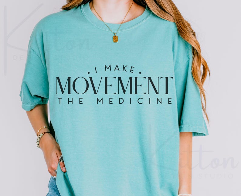 May include: A turquoise short-sleeved t-shirt with the text "I MAKE MOVEMENT THE MEDICINE" in black. The shirt has a crew neck and is worn by a person. A gold necklace and bracelets are also visible.