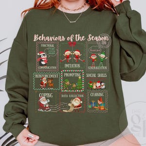 Könnte beinhalten: Ein olivgrünes Sweatshirt mit dem Text "Behaviors of the Season" und Illustrationen von weihnachtlichen Briefmarken. Die Briefmarken zeigen verschiedene Weihnachtsfiguren und Sätze wie "Communication" und "Imitation".