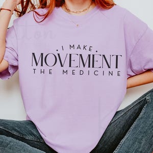 May include: A light purple t-shirt with the text "I MAKE MOVEMENT THE MEDICINE" in black. The shirt is a solid color and has short sleeves. The model is wearing blue jeans.