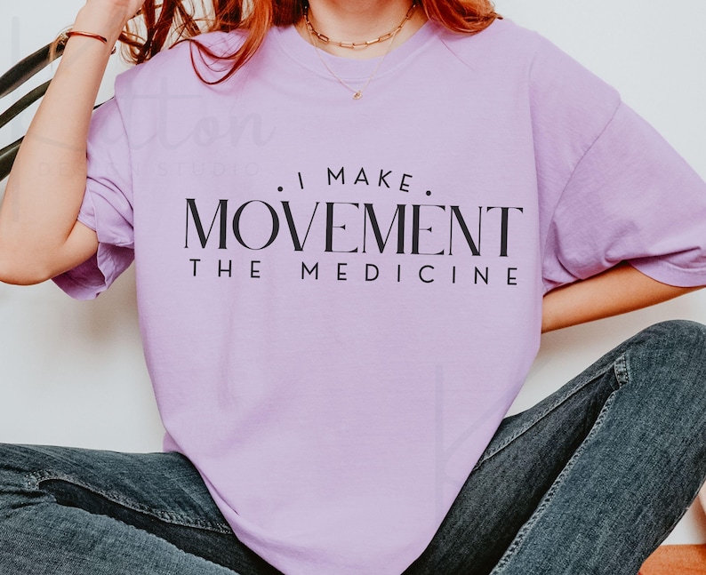 May include: A light purple t-shirt with the text "I MAKE MOVEMENT THE MEDICINE" in black. The shirt is a solid color and has short sleeves. The model is wearing blue jeans.