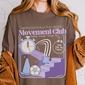 May include: Brown t-shirt with a graphic design featuring a stopwatch, stairs, a soccer ball, and the text "UNAPOLOGETICALLY PLAY-BASED Movement Club Building Strength Through Play." The design includes purple, blue, and white elements.