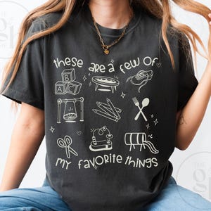 May include: Dark gray t-shirt with the phrase "these are a few of my favorite things" and various hand-drawn illustrations. The illustrations include ABC blocks, a swing, pencils, and a fork and spoon.